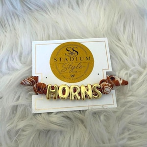 Puede incluir: Un coletero de tela con un patrón en color naranja quemado y blanco, adornado con cuentas de letras metálicas doradas que forman la palabra HORNS, sujeto a una tarjeta de presentación blanca con un logo circular dorado que dice STADIUM & STYLE CO.