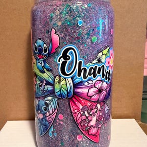 May include: A clear tumbler filled with purple glitter and a colorful bow design featuring the word "Ohana" and cartoon characters. The tumbler has a clear lid and is decorated with various images of the character Stitch.