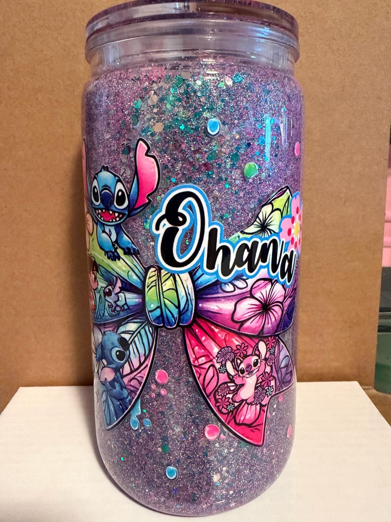 May include: A clear tumbler filled with purple glitter and a colorful bow design featuring the word "Ohana" and cartoon characters. The tumbler has a clear lid and is decorated with various images of the character Stitch.