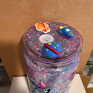May include: A clear tumbler with a purple glitter interior and a lid decorated with space-themed charms. The charms include a planet, a rocket, and a flying saucer. The tumbler has a cartoon character design on the side.