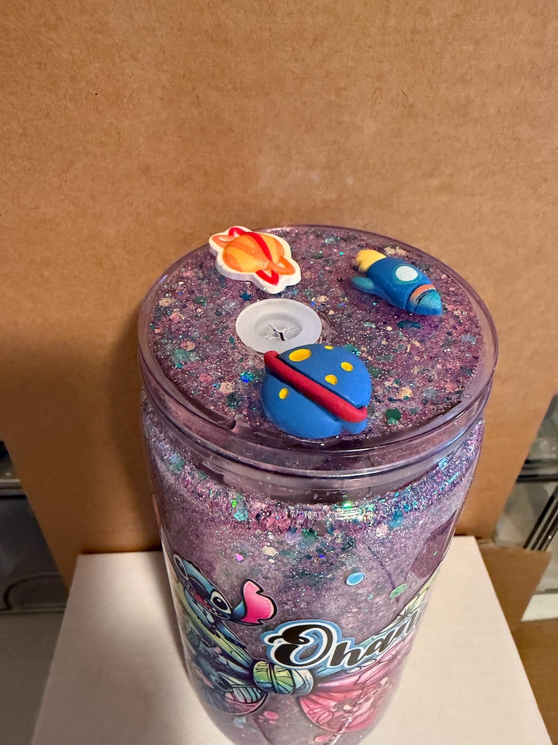 May include: A clear tumbler with a purple glitter interior and a lid decorated with space-themed charms. The charms include a planet, a rocket, and a flying saucer. The tumbler has a cartoon character design on the side.