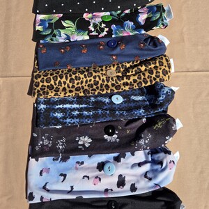 May include: A collection of eight fabric headbands arranged in a vertical stack. Each headband features a single central button and a unique pattern, including black with white polka dots, floral prints, leopard print, tie-dye, and solid black.