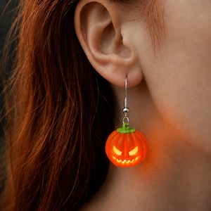 May include: A dangling earring featuring a small, bright orange jack-o'-lantern pumpkin charm with a green stem. The pumpkin has a carved, glowing face with triangular eyes and a jagged mouth, suspended from a silver-toned metal fishhook wire.