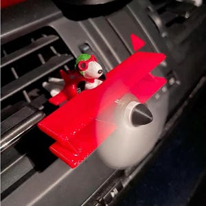 Puede incluir: Un ambientador de coche con forma de biplano rojo y un perro blanco con gorro de aviador verde y gafas rojas. El avión tiene un cono de nariz negro y una hélice giratoria, diseñado para fijarse en la rejilla de ventilación del vehículo.