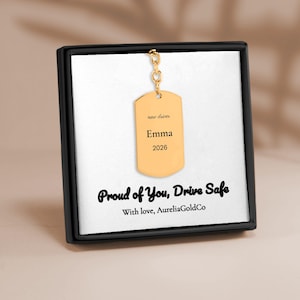 Peut inclure: Un porte-clés rectangulaire doré en forme de plaque militaire attaché à une chaîne, présenté dans une boîte noire. La plaque est gravée avec le texte new driver, Emma, et 2026. La carte blanche affiche Proud of You, Drive Safe et With love, AureliaGoldCo.