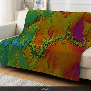 Puede incluir: Una manta suave con un diseño de mapa topográfico multicolor y vibrante. La superficie muestra un patrón en relieve con tonos verdes, naranjas, amarillos, azules y morados, que ilustra diversas elevaciones del terreno y ríos.