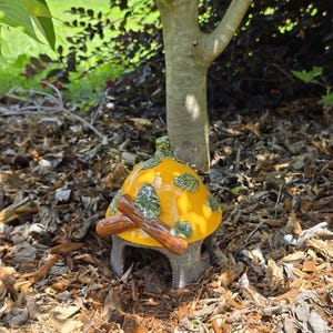 May include: A whimsical, yellow mushroom house with a brown roof and green leaf accents. The house has a small doorway and is decorated with two crossed brown logs. The house is set in a bed of mulch under a tree.