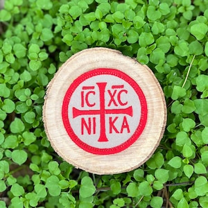 May include: A round wooden coaster with a red and gray embroidered patch. The patch features a red cross with Greek letters and the word "NIKA". The coaster is set against a backdrop of green foliage.