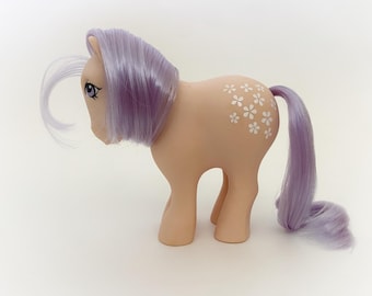 May include: A vintage toy pony in a standing pose. The pony has a peach-colored body with white flower designs on its flank. It features a lavender mane and tail, and a white forelock. The pony has blue eyes.