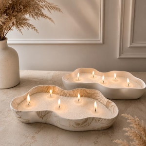 Peut inclure: Deux bols à bougies en céramique de forme irrégulière, remplis de cire blanche avec plusieurs mèches allumées. Le bol avant présente un motif marbré beige et blanc, tandis que le bol arrière est blanc uni. Ils contiennent des flammes vives.