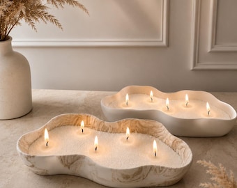 May include: Two irregular, cloud-shaped ceramic candle bowls filled with white wax and multiple lit wicks. The front bowl features a beige and white marbled pattern, while the back bowl is solid white. Both contain several small, bright flames.