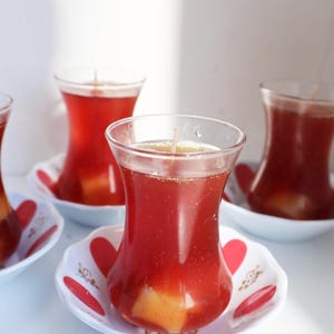 Può includere: Tre candele in bicchieri di vetro trasparente a forma di tulipano, riempite di cera rosso scuro con uno stoppino centrale. Le candele poggiano su piattini bianchi decorati con motivi a petali rossi e piccoli accenti dorati sui bordi.