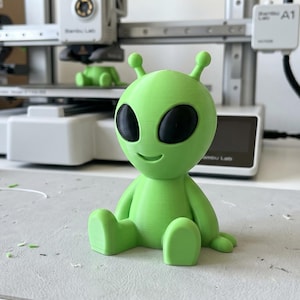 May include: A 3D printed lime green alien figurine with a smooth finish, large oval black eyes, and two antennae on its head. The alien is depicted in a seated position with its legs extended forward, showcasing a minimalist and clean design.