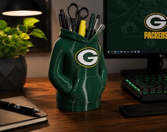 May include: A dark green desk organizer shaped like a hooded sweatshirt featuring the Green Bay Packers logo on the chest. The garment-style container has a front pouch pocket and an open hood at the top, currently holding various pens and scissors.