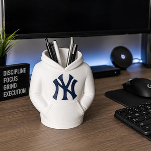 May include: A white desk organizer shaped like a hooded sweatshirt with a navy blue New York Yankees logo on the front. The container features a front pouch pocket design and holds several pens and pencils within its open top hood opening.