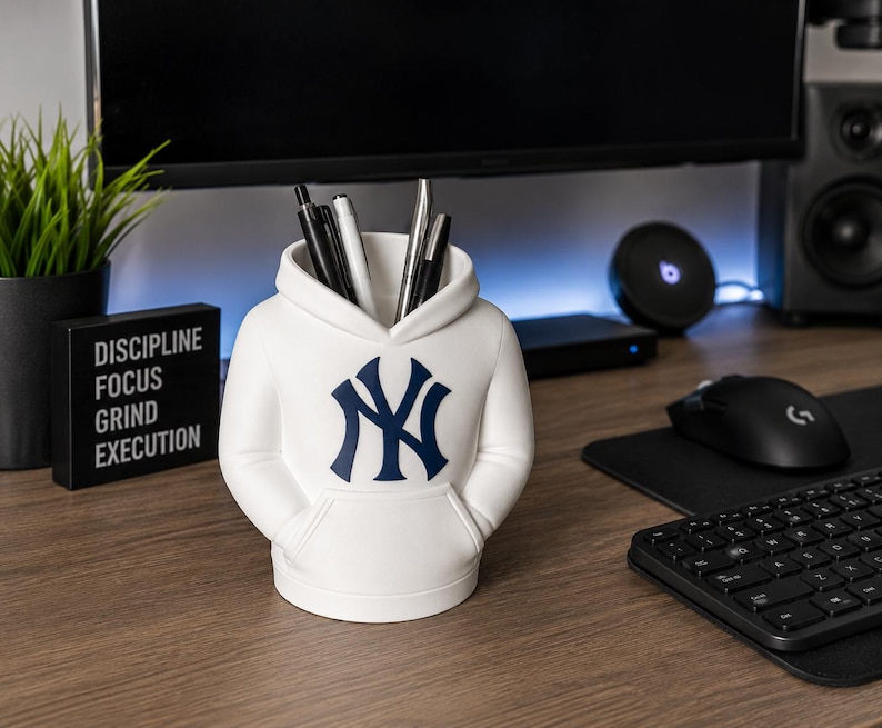 May include: A white desk organizer shaped like a hooded sweatshirt with a navy blue New York Yankees logo on the front. The container features a front pouch pocket design and holds several pens and pencils within its open top hood opening.