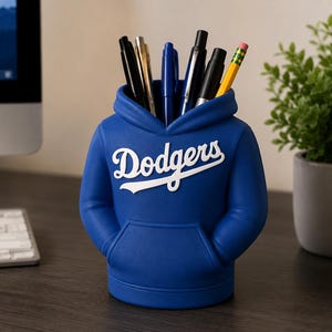 May include: A blue desk organizer shaped like a hooded sweatshirt with the word Dodgers printed in white script across the chest. The organizer features a front kangaroo pocket and holds several pens, pencils, and markers in the hood opening.