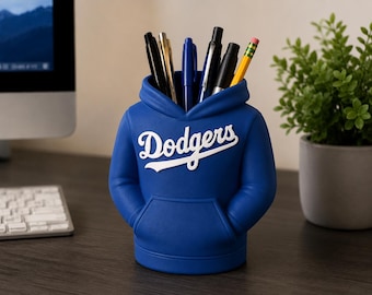 May include: A blue desk organizer shaped like a hooded sweatshirt with the word Dodgers printed in white script across the chest. The organizer features a front kangaroo pocket and holds several pens, pencils, and markers in the hood opening.
