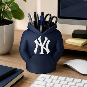 May include: A navy blue desk organizer shaped like a hooded sweatshirt featuring the white New York Yankees logo on the front. The organizer has a front pocket design and holds several pens, a silver mechanical pencil, and black scissors.