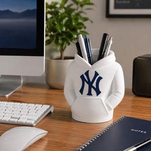 May include: A white desk organizer shaped like a hooded sweatshirt featuring the navy blue New York Yankees logo on the front. The container holds several pens and pencils, with a front pocket detail and a hood-shaped opening at the top.
