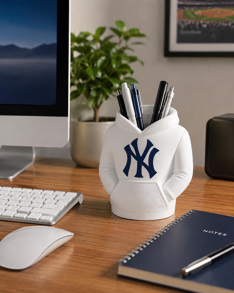 May include: A white desk organizer shaped like a hooded sweatshirt featuring the navy blue New York Yankees logo on the front. The container holds several pens and pencils, with a front pocket detail and a hood-shaped opening at the top.