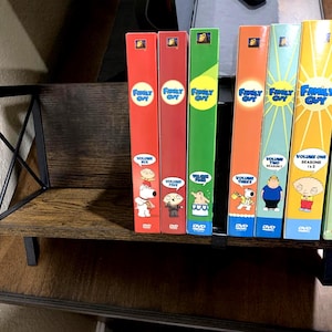 May include: A collection of Family Guy DVD box sets, featuring various volumes and seasons. The spines are in red, green, orange, and blue, with character illustrations. The DVDs are displayed on a dark wooden shelf with a black metal frame.