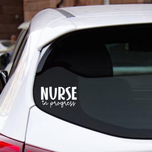 May include: White vinyl decal on a car window. The decal features the word "NURSE" in a bold font, with a heart symbol above the words "in progress" in a cursive script. The car is white.
