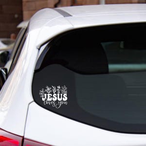 Può includere: Un adesivo in vinile bianco applicato sul lunotto posteriore di un veicolo bianco. L'adesivo presenta il testo 'JESUS loves you' in una combinazione di caratteri in grassetto e corsivo, circondato da varie illustrazioni di foglie e steli.