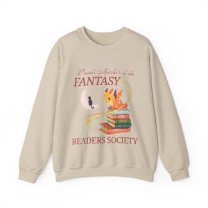 May include: Beige sweatshirt with a fantasy design. The graphic includes a stack of books, an orange dragon, a black cat on a crescent moon, and the text "Proud Member of the FANTASY READERS SOCIETY" in burgundy.
