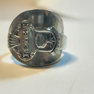 Puede incluir: Un anillo de metal plateado hecho a partir del mango de una cuchara, con el texto en relieve ROLEX y BUCHERER junto a un emblema de corona. El diseño incorpora los elementos decorativos originales del utensilio en una banda.
