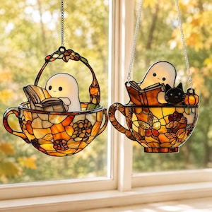 Op de afbeelding: Twee glas-in-lood zonnevangers in de vorm van theekopjes met oranje, gele en bruine bloemmotieven. Elk kopje bevat een wit spookfiguurtje dat een open boek leest. Eén kopje bevat een kleine zwarte kat en een kleine oranje pompoen.