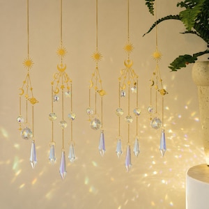 Può includere: Cinque ornamenti pendenti con ciondoli in metallo dorato a forma di sole, luna, stelle e pianeti sospesi su catenelle sottili. Ogni ornamento include cristalli di vetro sfaccettati trasparenti e prismi a goccia che riflettono la luce.