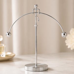 Puede incluir: Una escultura cinética de equilibrio de metal plateado con una varilla vertical central sobre una base circular. Un arco curvado se equilibra en la parte superior, con una pequeña figura estilizada en el vértice y una esfera de metal pulido en cada extremo.