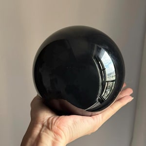 May include: A glossy, black sphere with a smooth, reflective surface. The sphere is held in a hand, and the surface reflects the surrounding environment. The object appears to be made of a solid, polished material.