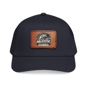 Puede incluir: Una gorra de béisbol azul marino con un parche rectangular de cuero marrón en la parte delantera. El parche muestra un gráfico en blanco y negro de un pez saltando fuera del agua con el texto MEATSTIK OUTDOORS HOOKED FOR LIFE impreso.