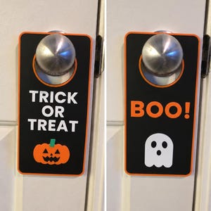 May include: A rectangular black door hanger with an orange border. One side features the white text TRICK OR TREAT above an orange jack-o'-lantern. The other side displays the orange text BOO! above a white ghost with two black eyes and a mouth.