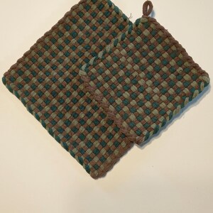 May include: Two square potholders featuring a woven checkerboard pattern in shades of teal, sage green, and brown. Each potholder has a thick braided border and a small fabric loop at one corner for hanging, constructed from soft textile material.