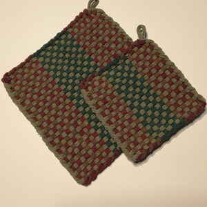 May include: Two square potholders featuring a woven pattern in shades of dark green, maroon, and tan. Each potholder has a small fabric loop attached to one corner for hanging. The woven texture is consistent across both items, which vary in size.