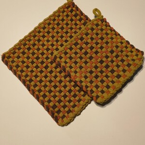 May include: Two square potholders featuring a woven checkerboard pattern in shades of dark brown, burnt orange, and mustard yellow. Each potholder is constructed from thick fabric loops and includes a small fabric loop at one corner for hanging.