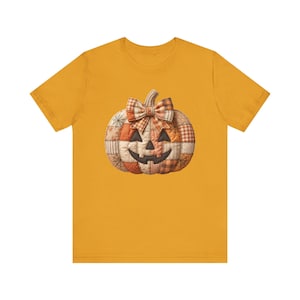 May include: A mustard yellow short-sleeve t-shirt featuring a central graphic of a jack-o'-lantern pumpkin. The pumpkin has a patchwork design with various orange, brown, and cream patterns, a plaid bow on top, and a dark brown stitched face.
