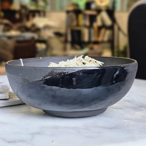 May include: A round, deep ceramic bowl with a matte dark gray exterior and a glossy black glazed rim. The bowl features a wide, open top and a sturdy base, containing a small portion of shredded white food material inside the basin.