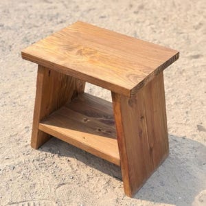 May include: A small wooden stool with a rectangular top and a lower shelf. The piece is constructed from medium-brown wood with a visible grain pattern, featuring two slanted side supports that connect the top surface to the base shelf.