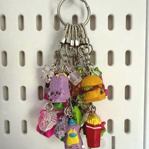 May include: A silver metal keyring featuring a cluster of colourful miniature plastic charms, including a purple bottle, a yellow hamburger, a red container of french fries, and various translucent beads attached with small silver lobster clasps.