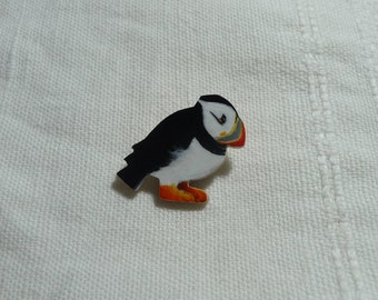 May include: A wooden pin of a black and white puffin with orange feet and a black beak. The puffin is facing to the right and is standing on a white surface.