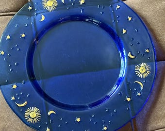 May include: A circular cobalt blue glass plate featuring a decorative rim adorned with gold-colored celestial motifs, including multiple suns, crescent moons, and various sizes of stars scattered across the surface of the plate's border.