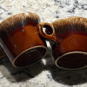 May include: Two ceramic mugs featuring a glossy brown glaze with a distinctive cream-colored drip pattern around the upper rim. Each mug has a sturdy handle and a circular base with embossed text visible on the bottom of the ceramic vessels.