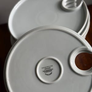 May include: Stack of white ceramic plates with a unique design. Each plate has a circular indentation and a curved handle-like feature. The plates are labeled "GREAT WHITE" and "MICROWAVE & DISHWASHER SAFE".