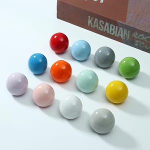 May include: A collection of twelve smooth, spherical knobs in various solid colors including red, light blue, dark gray, lime green, orange, mint green, yellow, lavender, pale pink, white, and light gray, arranged in a grid-like formation.