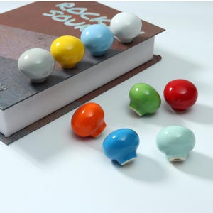Può includere: Una collezione di nove pomelli per armadietti lisci, rotondi e a forma di fungo in una varietà di colori lucidi tra cui bianco, azzurro, giallo, grigio, arancione, verde, rosso, blu scuro e turchese pallido, con una base piatta centrale.