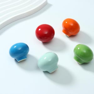May include: Five mushroom-shaped ceramic knobs in a glossy finish. The set includes one bright red, one vibrant orange, one light blue, one pale mint green, and one lime green knob. Each piece features a smooth, rounded cap and a small base.